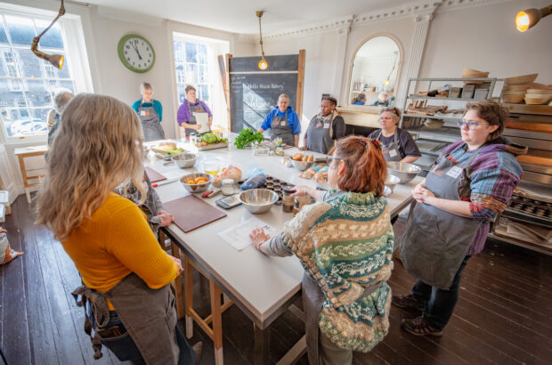 The group is standing around a large white table in the middle of the room. They're listening as they're told what recipes they will be making.